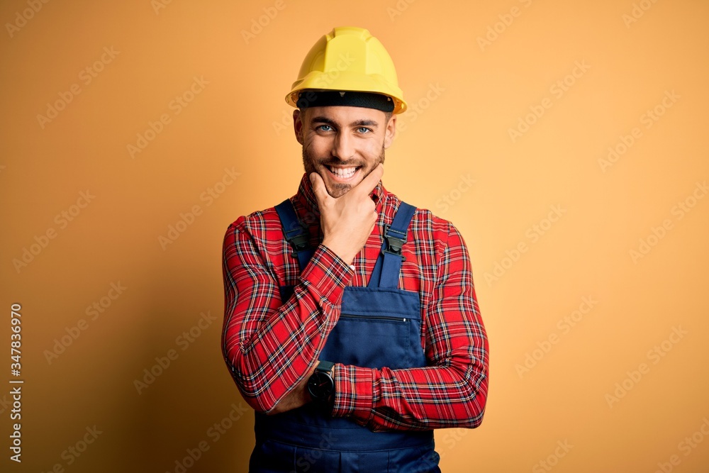 Young builder man wearing construction uniform and safety helmet over yellow isolated background looking confident at the camera smiling with crossed arms and hand raised on chin. Thinking positive.