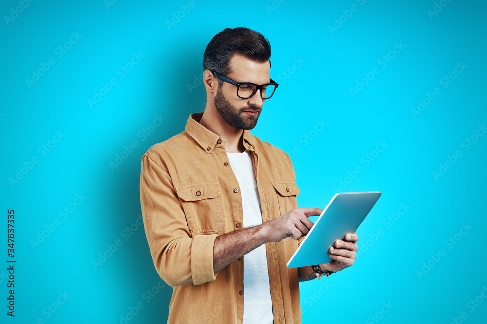 Busy young man in smart casual wear using digital tablet while standing against blue background Busy young man in smart casual wear using digital tablet while standing against blue background