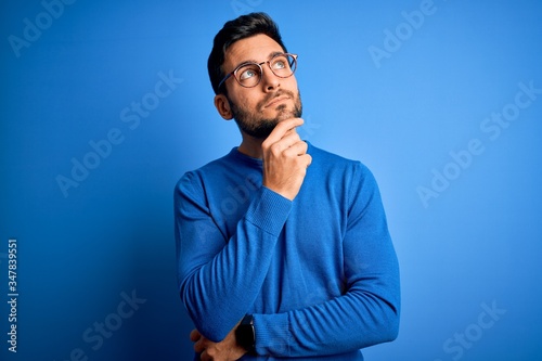Young handsome man with beard wearing casual sweater and glasses over blue background with hand on chin thinking about question, pensive expression. Smiling with thoughtful face. Doubt concept.