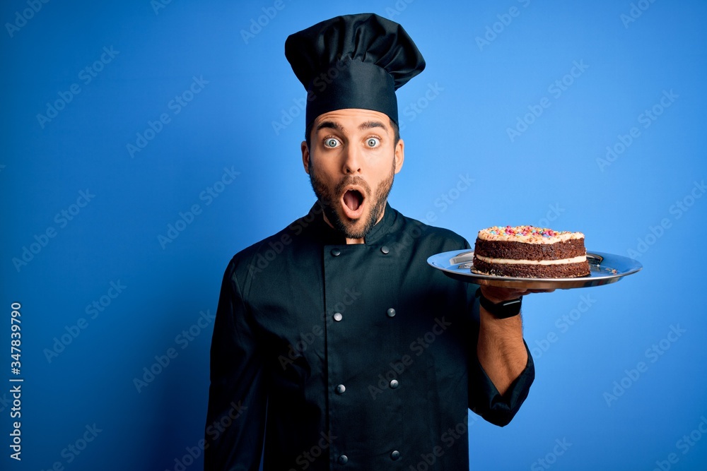 Young handsome cooker man with beard wearing uniform and hat holding tray with cake scared in shock with a surprise face, afraid and excited with fear expression Young handsome cooker man with beard wearing uniform and hat holding tray with cake scared in shock with a surprise face, afraid and excited with fear expression
