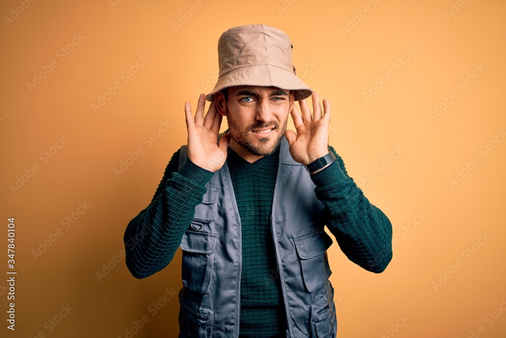 Handsome tourist man with beard on vacation wearing explorer hat over yellow background Trying to hear both hands on ear gesture, curious for gossip. Hearing problem, deaf Handsome tourist man with beard on vacation wearing explorer hat over yellow background Trying to hear both hands on ear gesture, curious for gossip. Hearing problem, deaf