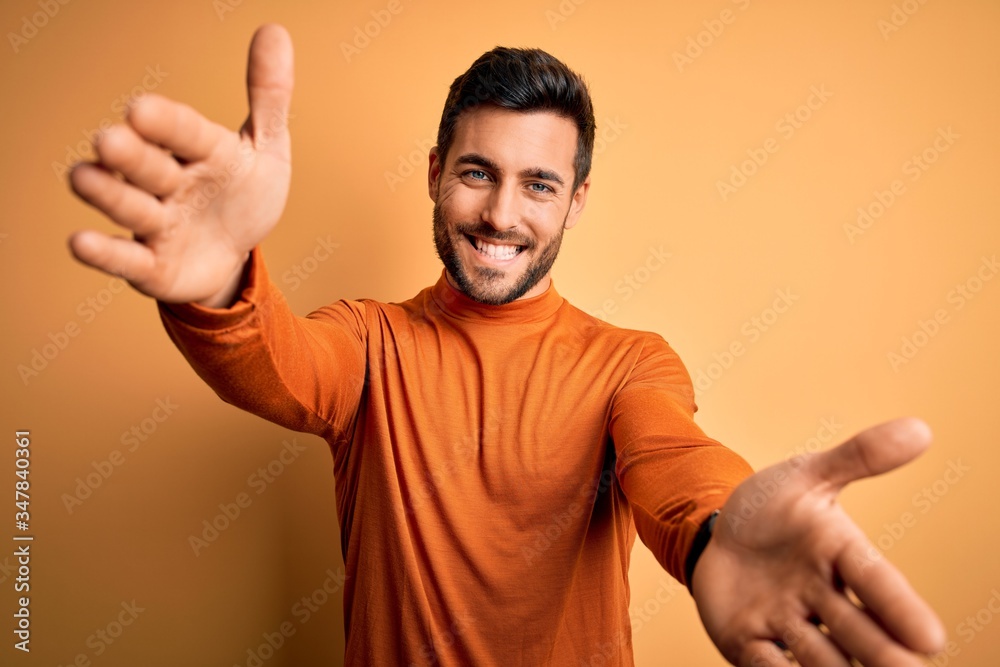 Young handsome man with beard wearing casual sweater standing over yellow background looking at the camera smiling with open arms for hug. Cheerful expression embracing happiness. Young handsome man with beard wearing casual sweater standing over yellow background looking at the camera smiling with open arms for hug. Cheerful expression embracing happiness.