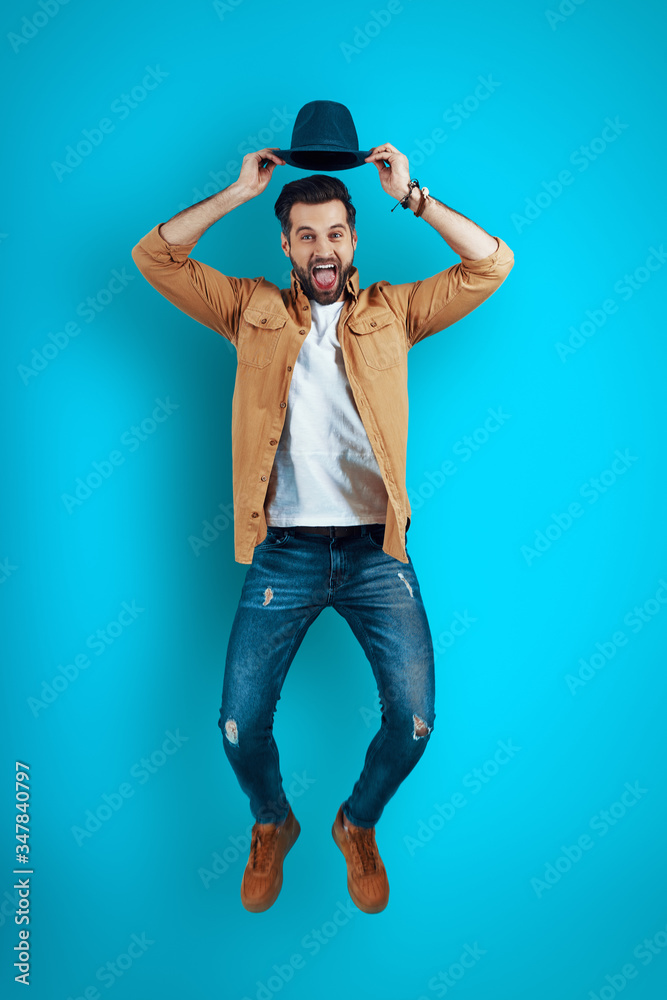 Full length of handsome young man smiling and adjusting hat while hovering against blue background Full length of handsome young man smiling and adjusting hat while hovering against blue background