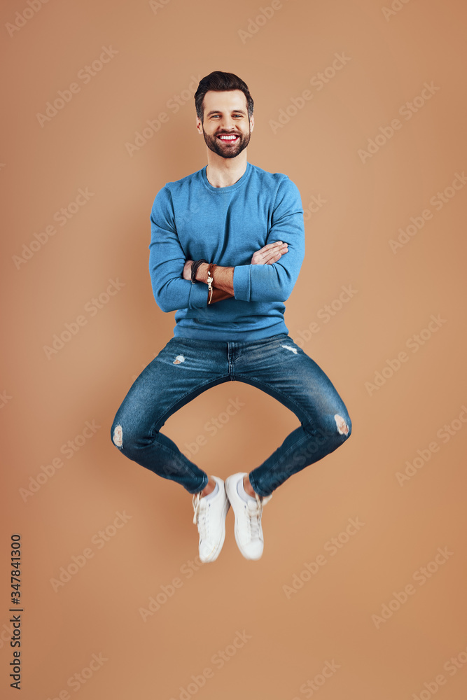Full length of happy young man smiling and keeping arms crossed while hovering against brown background Full length of happy young man smiling and keeping arms crossed while hovering against brown background