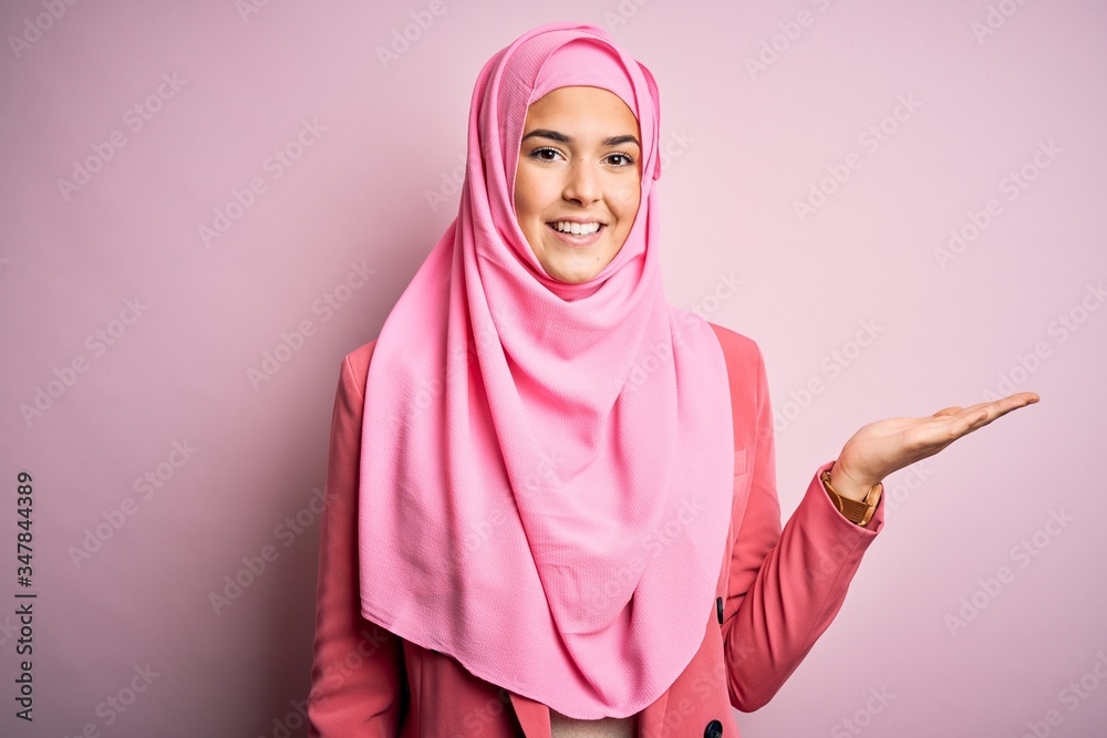 Young beautiful girl wearing muslim hijab standing over isolated pink background smiling cheerful presenting and pointing with palm of hand looking at the camera. Young beautiful girl wearing muslim hijab standing over isolated pink background smiling cheerful presenting and pointing with palm of hand looking at the camera.