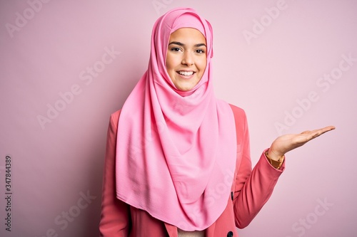 Young beautiful girl wearing muslim hijab standing over isolated pink background smiling cheerful presenting and pointing with palm of hand looking at the camera. © Krakenimages.com