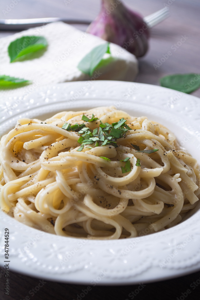 A vegan version of traditional Italian pasta fettuccine alfredo with creamy white sauce garnished with basil on a wooden surface with a fork, napkin, garlic and basil leaves A vegan version of traditional Italian pasta fettuccine alfredo with creamy white sauce garnished with basil on a wooden surface with a fork, napkin, garlic and basil leaves