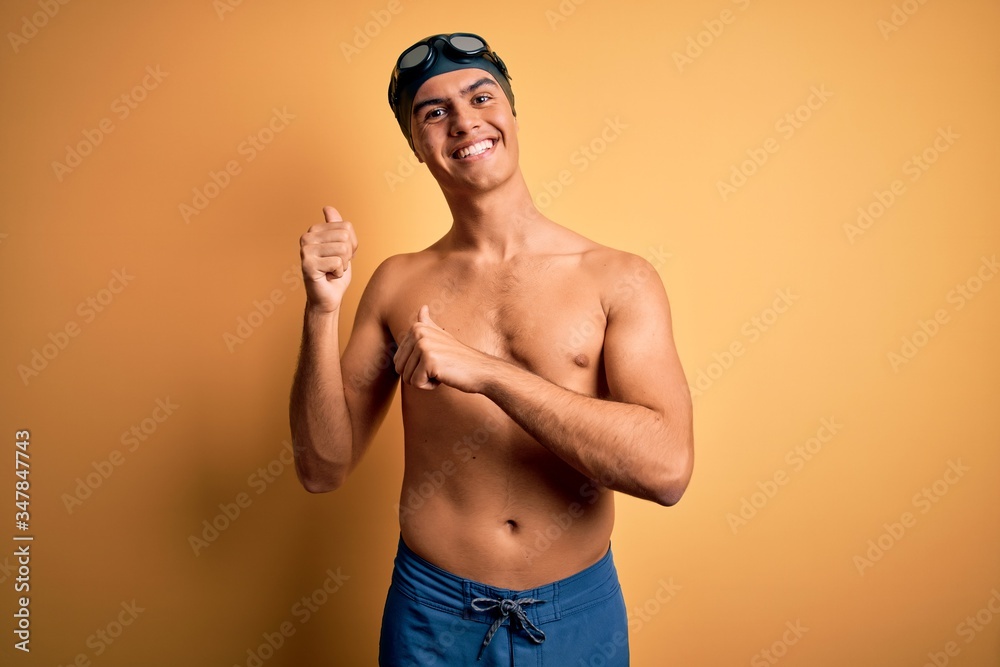 Young handsome man shirtless wearing swimsuit and swim cap over isolated yellow background Pointing to the back behind with hand and thumbs up, smiling confident