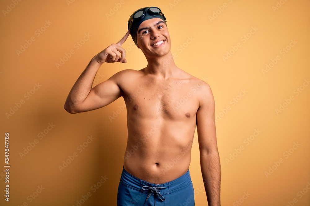 Young handsome man shirtless wearing swimsuit and swim cap over isolated yellow background Smiling pointing to head with one finger, great idea or thought, good memory