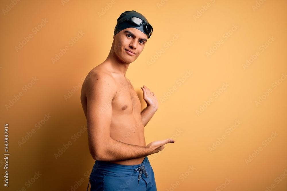 Young handsome man shirtless wearing swimsuit and swim cap over isolated yellow background Inviting to enter smiling natural with open hand