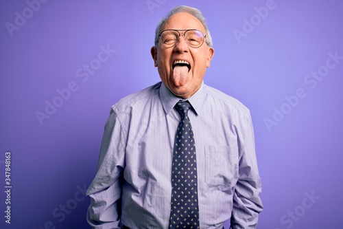 Grey haired senior business man wearing glasses standing over purple isolated background sticking tongue out happy with funny expression. Emotion concept.