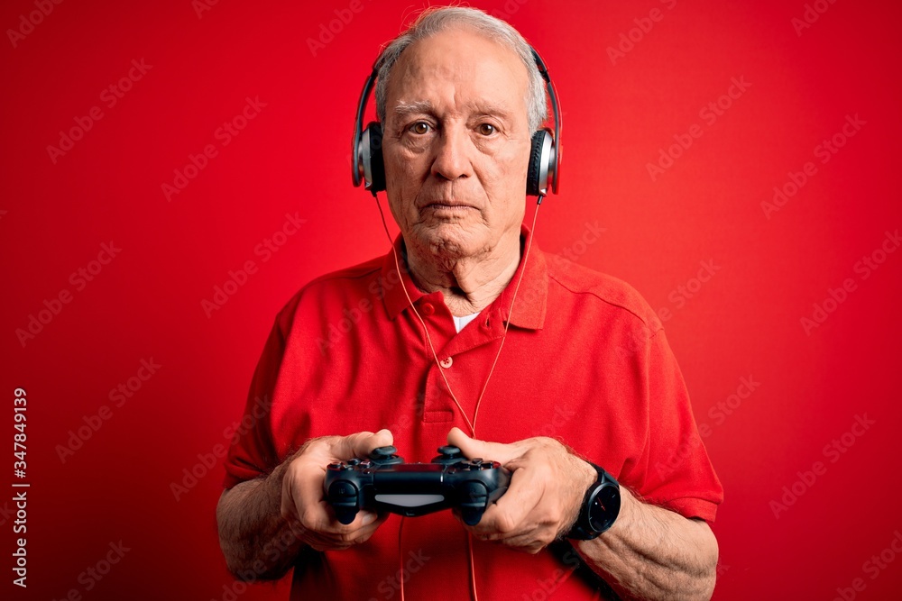 Senior grey haired gamer man playing video games using gamepad joystick over red background with a confident expression on smart face thinking serious Senior grey haired gamer man playing video games using gamepad joystick over red background with a confident expression on smart face thinking serious