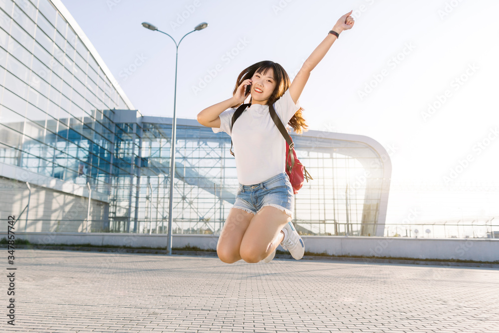 Funny photo of beautiful excited young asian woman in jeans shorts and white t-shirt, with red backpack, talking phone, and jumping up with one hand raised, waiting for journey near modern airport
