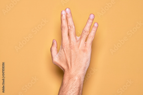 Hand of caucasian young man showing fingers over isolated yellow background greeting doing Vulcan salute  showing back of the hand and fingers  freak culture