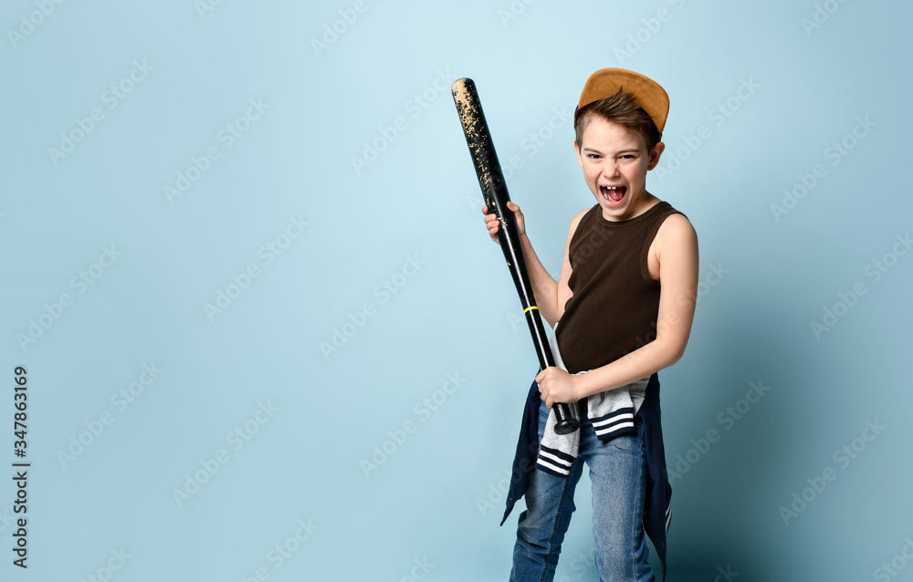 Teenage child in cap and casual clothes. Holding black baseball bat or club and screaming, posing against blue studio background Teenage child in cap and casual clothes. Holding black baseball bat or club and screaming, posing against blue studio background