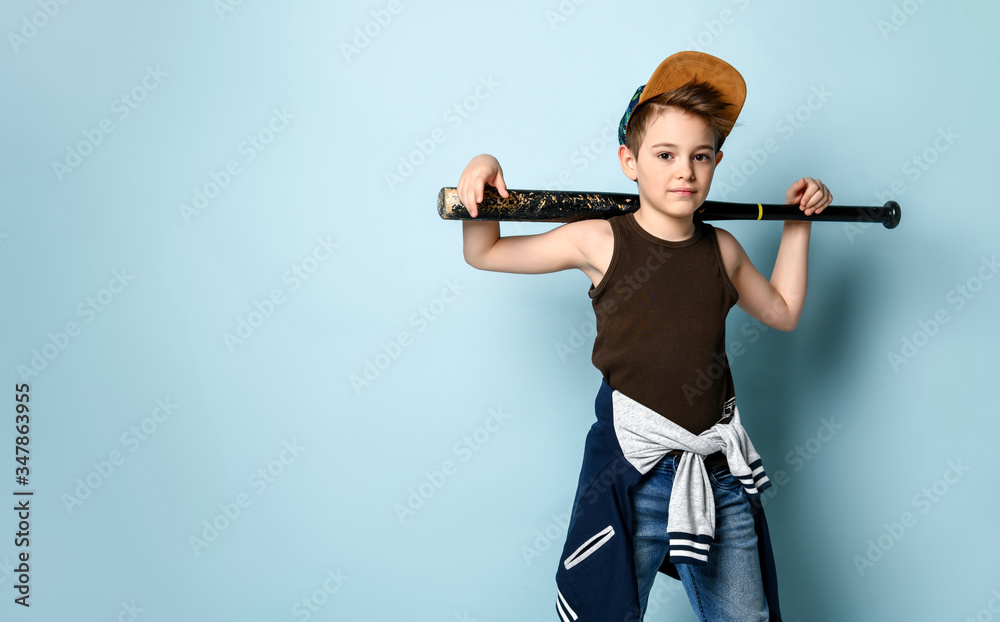 Teenage boy in cap, undershirt, jeans and hoodie tied around his waist. Holding black baseball bat, posing on blue background Teenage boy in cap, undershirt, jeans and hoodie tied around his waist. Holding black baseball bat, posing on blue background