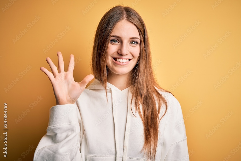 Young beautiful redhead sporty woman wearing sweatshirt over isolated yellow background showing and pointing up with fingers number five while smiling confident and happy. Young beautiful redhead sporty woman wearing sweatshirt over isolated yellow background showing and pointing up with fingers number five while smiling confident and happy.