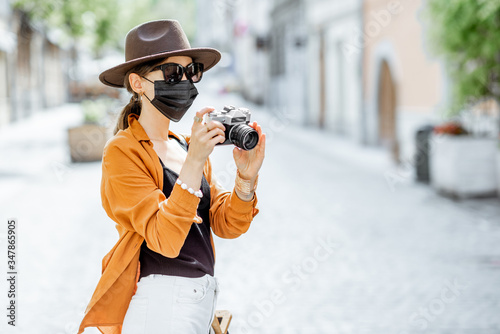 Young female tourist in facial protective mask walking with photo camera on the old city street. Concept of tourism and new social rules and restrictions after coronavirus pandemic