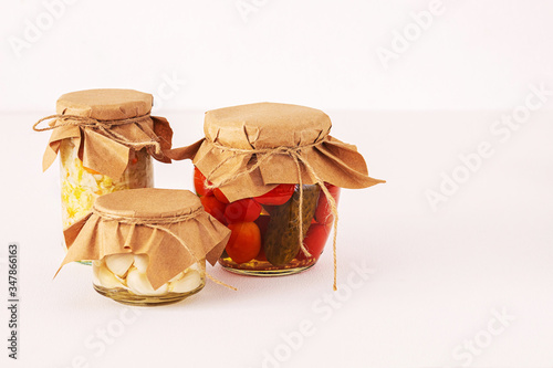 fermented vegetables in transparent glass jars covered with paper from recycled materials. tomatoes, garlic, cabbage. On a white pinkish background.