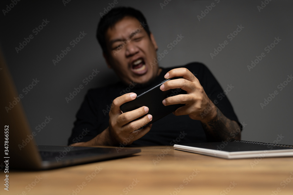 Asian man playing games with cellphone on wooden table with laptop. with funny face. Asian man playing games with cellphone on wooden table with laptop. with funny face.