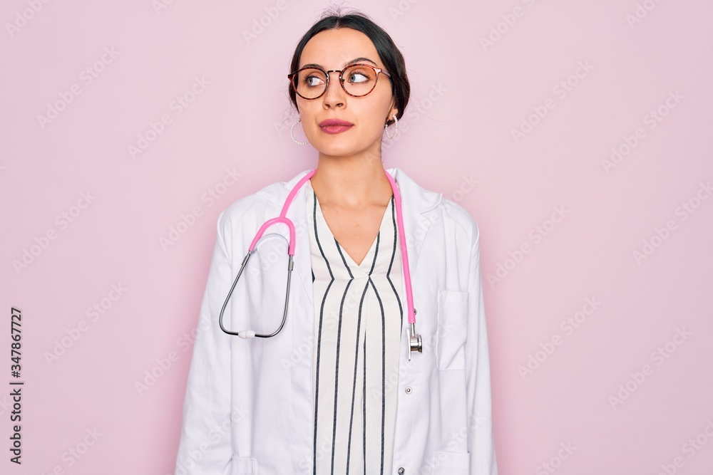 Beautiful doctor woman with blue eyes wearing coat and stethoscope over pink background smiling looking to the side and staring away thinking. Beautiful doctor woman with blue eyes wearing coat and stethoscope over pink background smiling looking to the side and staring away thinking.