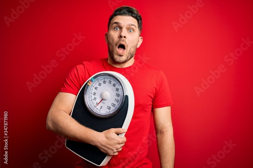 Young fitness man with blue eyes holding scale dieting for healthy weight over red background scared in shock with a surprise face, afraid and excited with fear expression