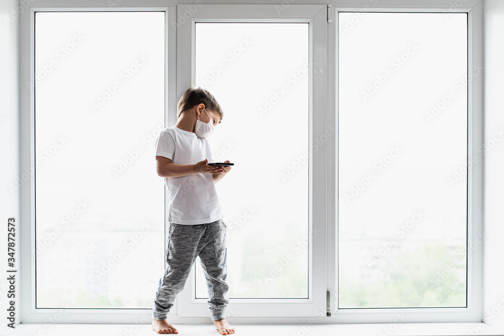 A little boy in a medical mask is sitting on the window of the house in quarantine with a phone in his hands.Prevention of coronavirus and Covid - 19 A little boy in a medical mask is sitting on the window of the house in quarantine with a phone in his hands.Prevention of coronavirus and Covid - 19