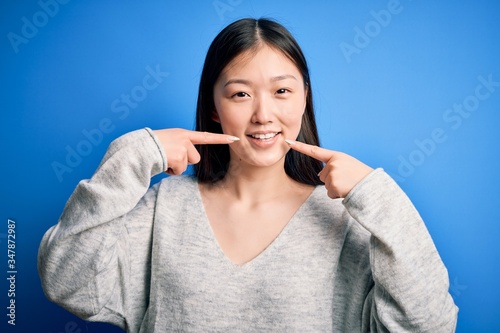 Young beautiful asian woman wearing casual sweater standing over blue isolated background smiling cheerful showing and pointing with fingers teeth and mouth. Dental health concept.
