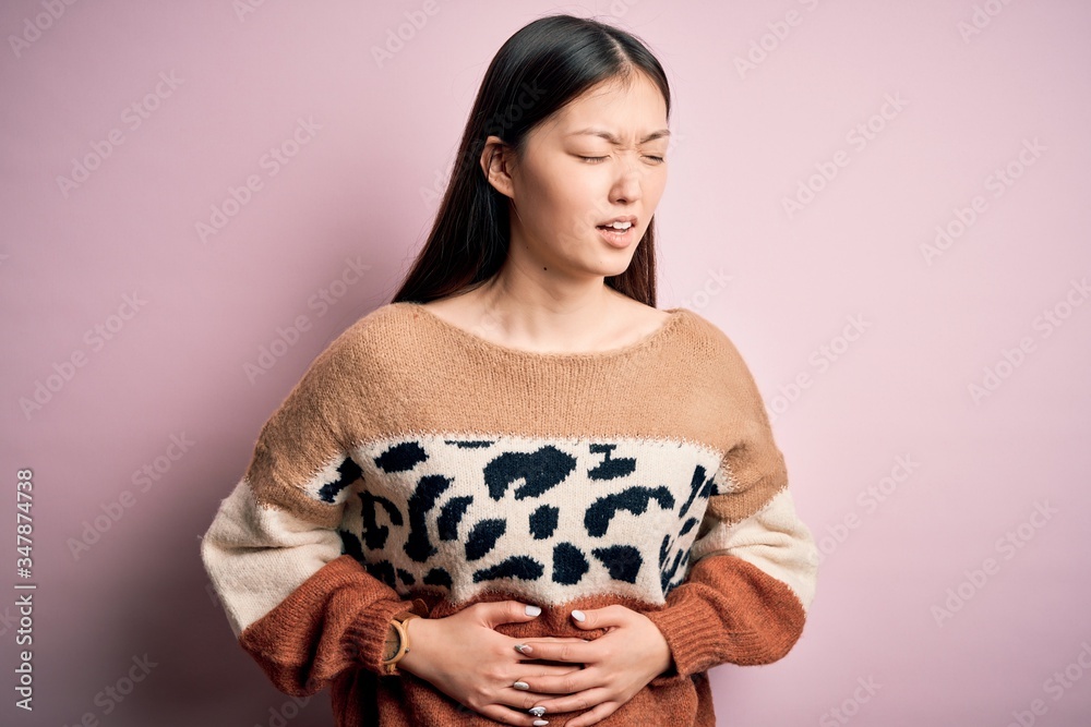 Young beautiful asian woman wearing animal print fashion sweater over pink isolated background with hand on stomach because nausea, painful disease feeling unwell. Ache concept. Young beautiful asian woman wearing animal print fashion sweater over pink isolated background with hand on stomach because nausea, painful disease feeling unwell. Ache concept.