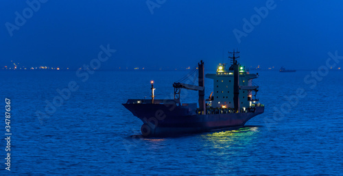 Container cargo ship with navigation lights anchored in outer anchorage of Singapore at golden hour.