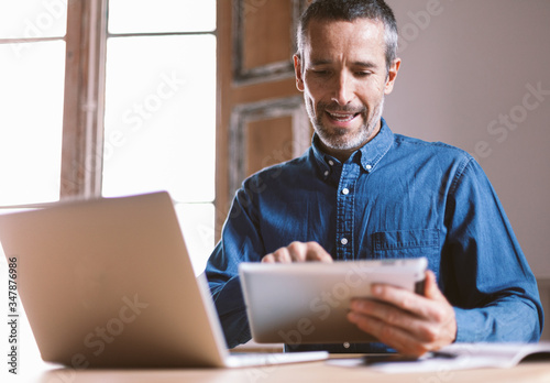Good looking middle aged modern man having an online meeting on his laptop and tablet from home with his colleagues