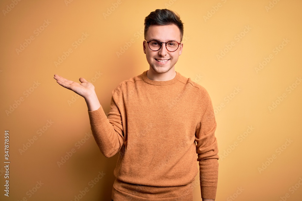 Young handsome caucasian man wearing glasses and casual winter sweater over yellow background smiling cheerful presenting and pointing with palm of hand looking at the camera. Young handsome caucasian man wearing glasses and casual winter sweater over yellow background smiling cheerful presenting and pointing with palm of hand looking at the camera.
