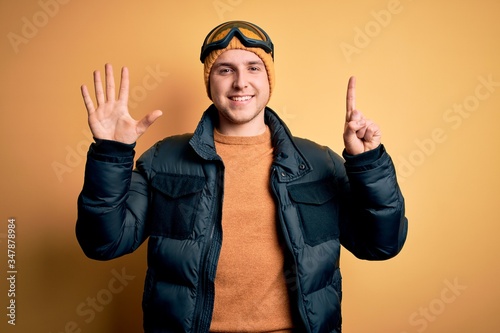 Young handsome caucasian man wearing hat, coat and ski glasses for winter and snow weather showing and pointing up with fingers number six while smiling confident and happy.