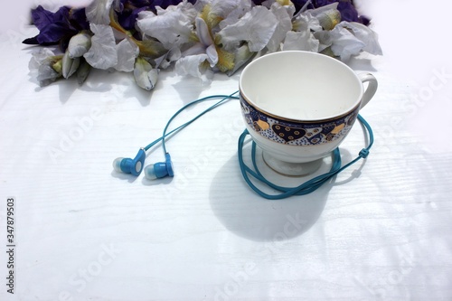 Obraz na płótnie Blue flowers with headphones in a white porcelain cup with a blue-gold engraving. On a white background, a porcelain cup with a blue iris flower and headphones. w salonie