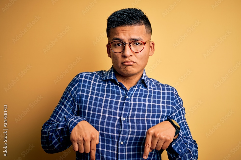 Young handsome latin man wearing casual shirt and glasses over yellow background Pointing down looking sad and upset, indicating direction with fingers, unhappy and depressed. Young handsome latin man wearing casual shirt and glasses over yellow background Pointing down looking sad and upset, indicating direction with fingers, unhappy and depressed.