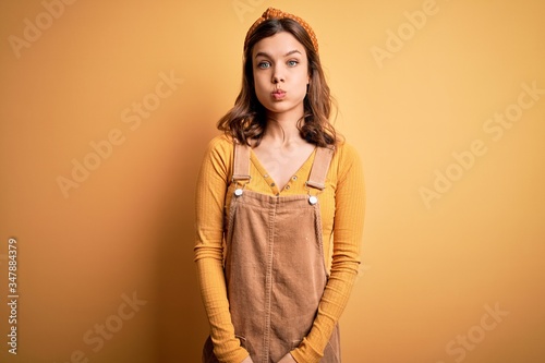 Young beautiful blonde girl wearing overall standing over yellow isolated background puffing cheeks with funny face. Mouth inflated with air, crazy expression.