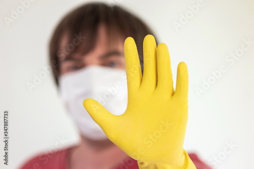 Out of focus Caucasian male with medical mask covering his face holding out his right hand with yellow latex glove in a STOP sign gesture