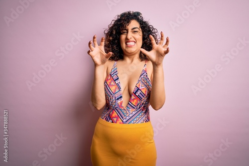 Young beautiful arab woman on vacation wearing swimsuit and sunglasses over pink background smiling funny doing claw gesture as cat  aggressive and sexy expression