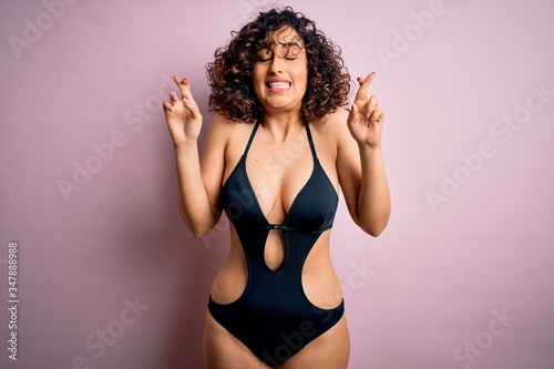 Young beautiful arab woman on vacation wearing swimsuit and sunglasses over pink background gesturing finger crossed smiling with hope and eyes closed. Luck and superstitious concept.