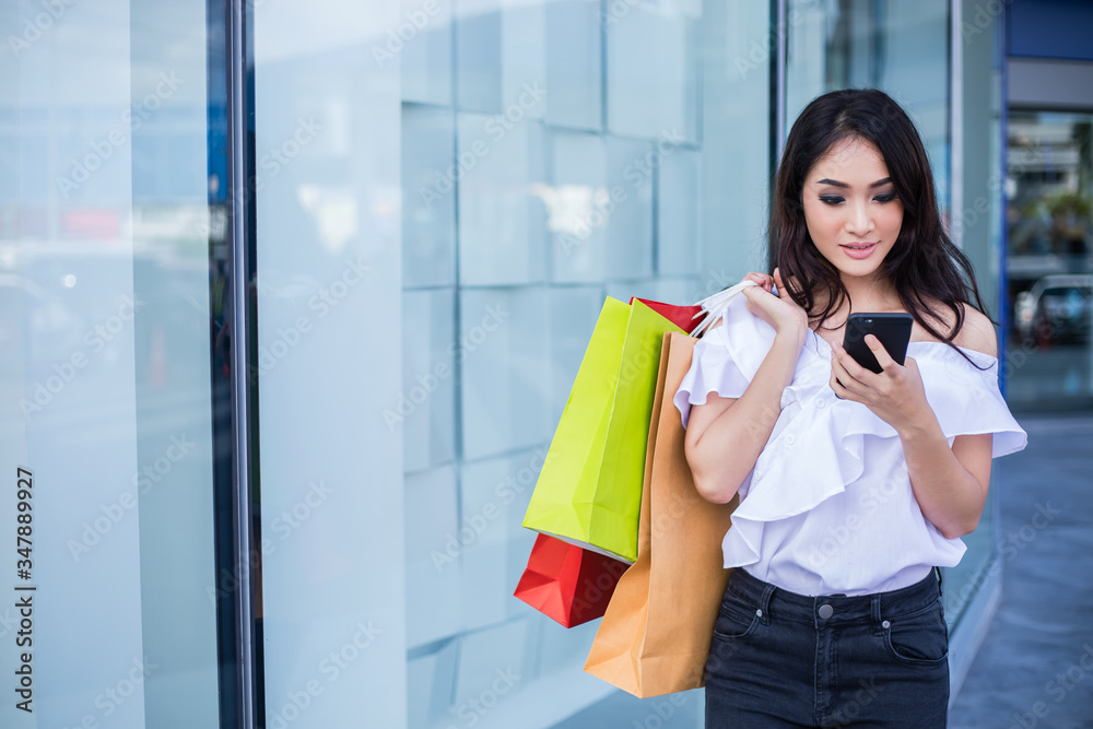 Beautiful asian young woman with shopping bags using her smart phone with smile while standing at the clothing store. Happiness, consumerism, sale and people concept Beautiful asian young woman with shopping bags using her smart phone with smile while standing at the clothing store. Happiness, consumerism, sale and people concept
