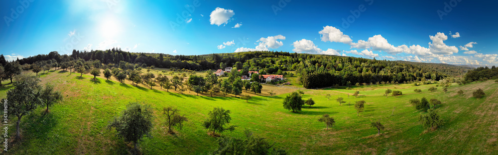Wide aerial panorama view of a nice rural landscape with green meadows on hills, trees and a few houses, deep blue sky, the sun and white clouds