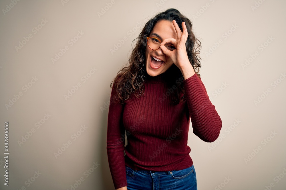 Beautiful woman with curly hair wearing casual sweater and glasses over white background doing ok gesture with hand smiling, eye looking through fingers with happy face. Beautiful woman with curly hair wearing casual sweater and glasses over white background doing ok gesture with hand smiling, eye looking through fingers with happy face.