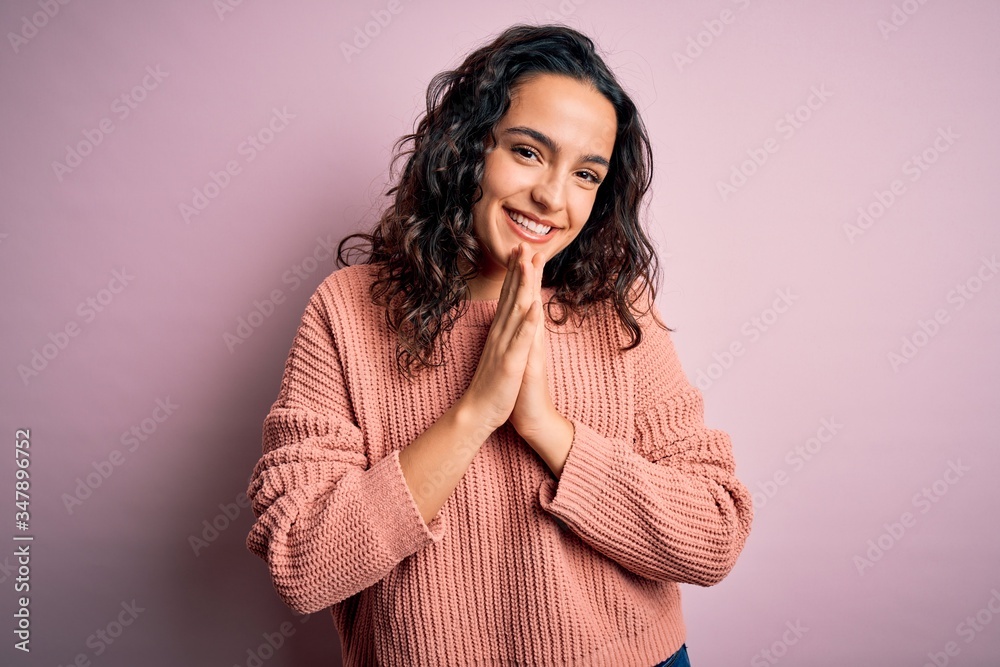 Young beautiful woman with curly hair wearing casual sweater over isolated pink background praying with hands together asking for forgiveness smiling confident. Young beautiful woman with curly hair wearing casual sweater over isolated pink background praying with hands together asking for forgiveness smiling confident.