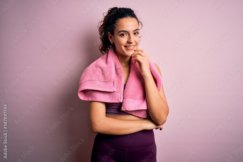 Young beautiful sportswoman with curly hair doing sport using towel over pink background looking confident at the camera with smile with crossed arms and hand raised on chin. Thinking positive. Young beautiful sportswoman with curly hair doing sport using towel over pink background looking confident at the camera with smile with crossed arms and hand raised on chin. Thinking positive.