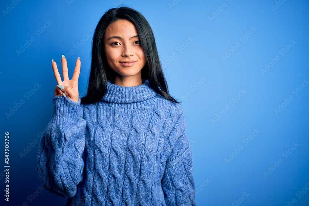 Young beautiful chinese woman wearing casual sweater over isolated blue background showing and pointing up with fingers number three while smiling confident and happy. Young beautiful chinese woman wearing casual sweater over isolated blue background showing and pointing up with fingers number three while smiling confident and happy.
