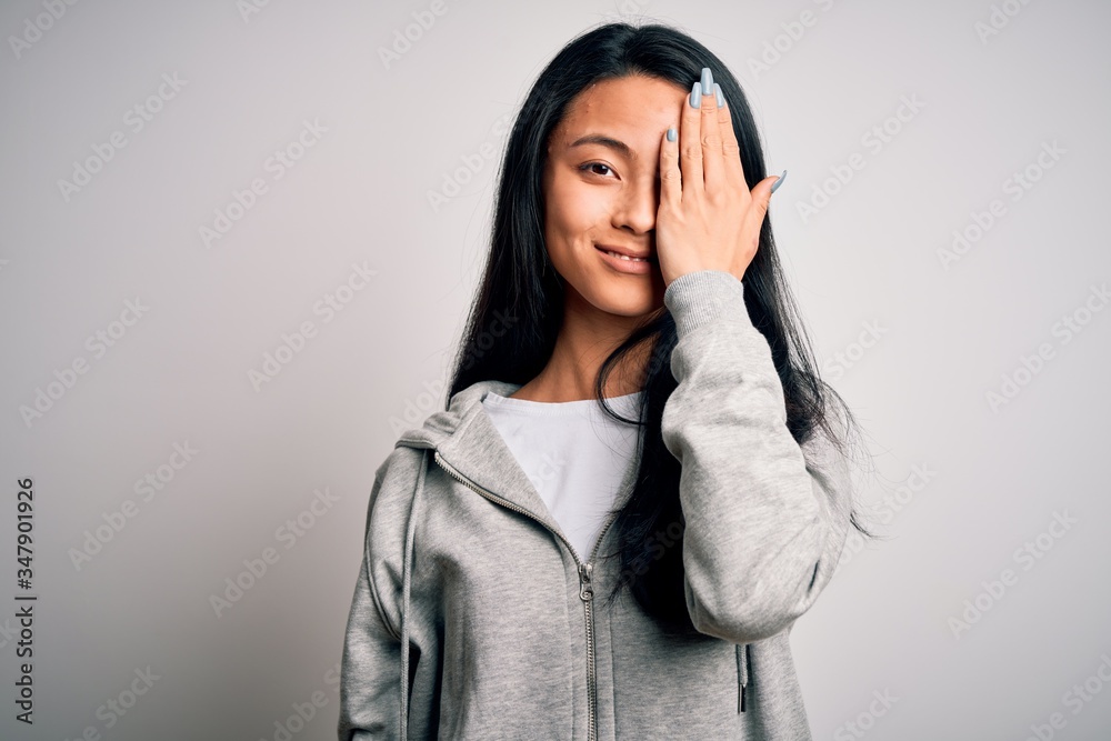 Young beautiful chinese sporty woman wearing sweatshirt over isolated white background covering one eye with hand, confident smile on face and surprise emotion. Young beautiful chinese sporty woman wearing sweatshirt over isolated white background covering one eye with hand, confident smile on face and surprise emotion.