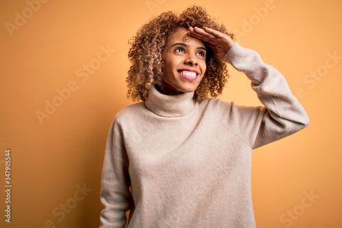 Young beautiful african american woman wearing turtleneck sweater over yellow background very happy and smiling looking far away with hand over head. Searching concept. © Krakenimages.com