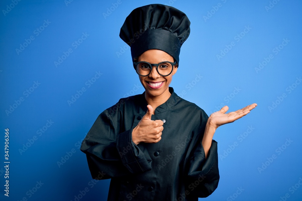 Young african american chef woman wearing cooker uniform and hat over blue background Showing palm hand and doing ok gesture with thumbs up, smiling happy and cheerful Young african american chef woman wearing cooker uniform and hat over blue background Showing palm hand and doing ok gesture with thumbs up, smiling happy and cheerful