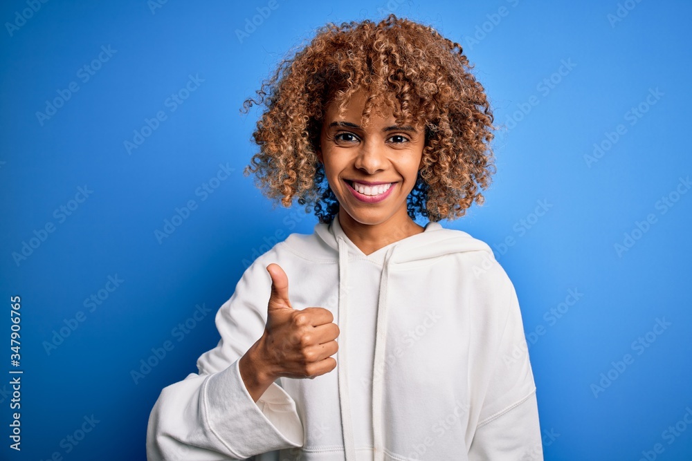 Beautiful african american sporty woman wearing casual sweatshirt over blue background doing happy thumbs up gesture with hand. Approving expression looking at the camera showing success. Beautiful african american sporty woman wearing casual sweatshirt over blue background doing happy thumbs up gesture with hand. Approving expression looking at the camera showing success.