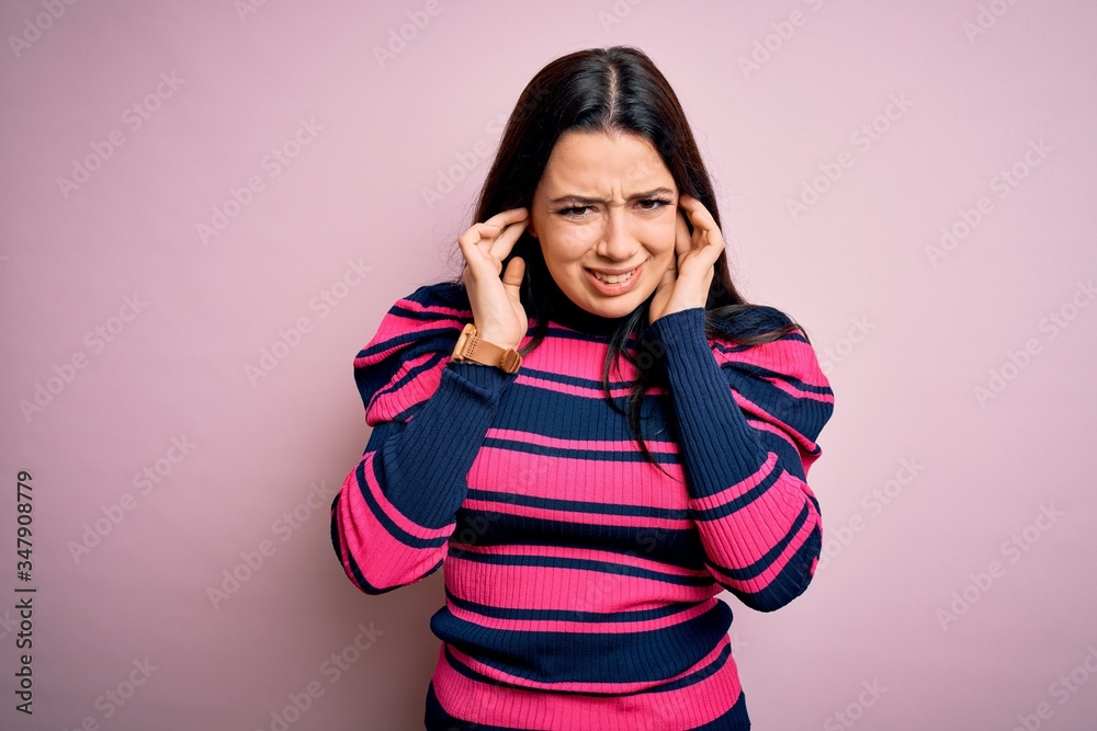 Young brunette elegant woman wearing striped shirt over pink isolated background covering ears with fingers with annoyed expression for the noise of loud music. Deaf concept. Young brunette elegant woman wearing striped shirt over pink isolated background covering ears with fingers with annoyed expression for the noise of loud music. Deaf concept.